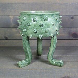 May include: A green ceramic planter with a textured surface and spiky protrusions. The planter is supported by three leg-like structures, giving it a unique and whimsical appearance. The planter is designed to hold plants and add a decorative touch to any space.