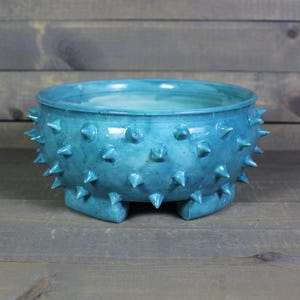 May include: A turquoise ceramic bowl with a textured surface and spiky protrusions around the exterior. The bowl has a wide, open top and sits on three small feet. The bowl is approximately 10 inches in diameter.