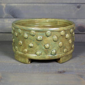 May include: A green ceramic planter with a textured, raised dot pattern around the middle. The planter has three short legs and a rounded base.