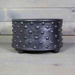 May include: A black ceramic planter with a textured surface and small, rounded studs. The planter has a low, wide shape and sits on three small feet. The background is a weathered wooden surface.