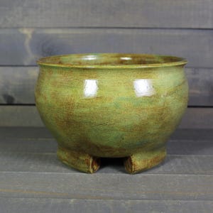 May include: A round, green ceramic bowl with three small feet. The bowl has a glossy finish with brown and green tones. The handmade pottery bowl is suitable for home decor or as a planter.