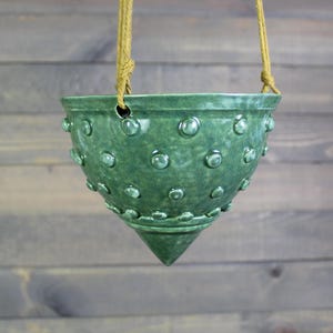 May include: A hanging green ceramic planter with a textured surface of raised, rounded bumps. The planter has a conical base and is suspended by a tan rope. The planter is designed for indoor or outdoor use.