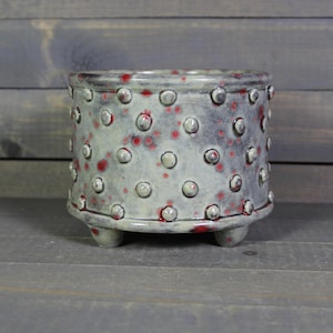 May include: A ceramic planter with a speckled, light gray glaze and red accents. The cylindrical pot features a pattern of raised, rounded dots and rests on three small, rounded feet. The interior and rim show a darker, reddish hue.