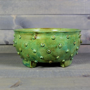 May include: A handcrafted, green ceramic bowl with a textured surface and three small feet. The bowl features a series of raised, circular designs across its exterior. The glaze has a mottled appearance, with variations in the green color.