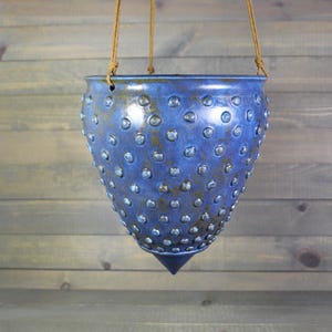 May include: A cone-shaped hanging planter with a vibrant blue glaze and a textured surface covered in small, raised circular studs. The planter features a pointed bottom and is suspended by three thin, light brown cords attached to the rim.