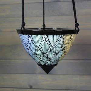 May include: A hanging ceramic planter with a black rim and base. The white body is decorated with a black geometric pattern of vertical lines and diamond shapes. Black rope suspends the planter, creating a modern, decorative piece.