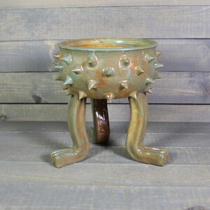 May include: A ceramic planter with a unique design. The planter is a mix of green and brown hues, featuring a textured surface with spike-like protrusions. It stands on three leg-like supports and a tail-like support.