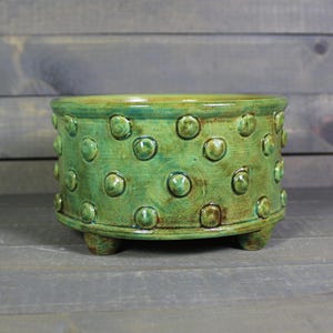 May include: A handcrafted, green ceramic bowl with a textured, dotted pattern. The bowl has a rounded shape and sits on three small feet. The glaze has a glossy finish with variations in color, adding depth and character to the piece.