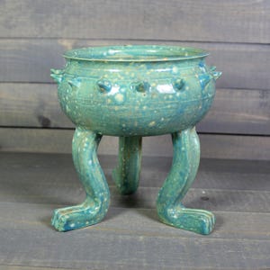 May include: A turquoise ceramic bowl with three leg-like supports. The bowl has a rounded shape with a textured surface and small, raised protrusions around the rim. The legs end in a foot-like shape. The piece has a speckled appearance.