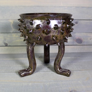 May include: A handcrafted, dark purple ceramic bowl with a textured surface and spiky protrusions. The bowl is supported by three curved legs, giving it a unique, artistic appearance. The interior has a dark, glossy finish.
