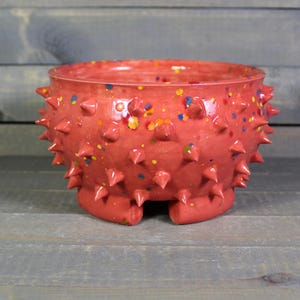 May include: A handmade, red ceramic bowl with a textured surface of conical spikes. The bowl is speckled with colorful paint flecks and has three small feet. This unique piece is ideal for home decor.