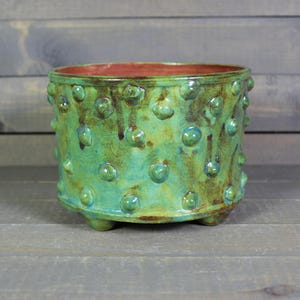 May include: A handcrafted ceramic planter with a textured, bumpy surface. The planter is glazed in shades of turquoise and green, with brown accents. It has a reddish-brown interior and sits on three small feet. A decorative piece for plants.