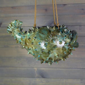 May include: A hanging ceramic pufferfish planter featuring a glossy, mottled green and blue glaze. The fish-shaped vessel is covered in numerous small, pointed spikes and includes a side fin, a tail, and two brown cords for suspension.