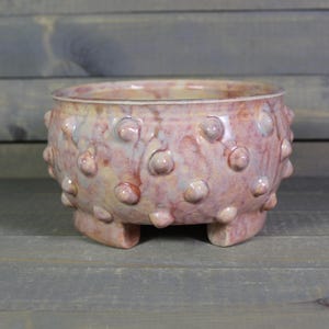 May include: A handcrafted ceramic bowl with a textured surface and a unique design. The bowl is a blend of pink, cream, and lavender hues, with rounded protrusions around the exterior. It rests on three small feet.