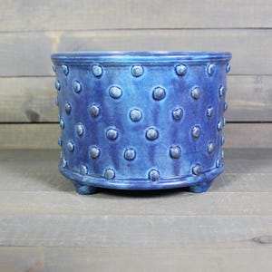 May include: A cylindrical ceramic planter with a deep blue glazed finish and a repeating pattern of raised, circular dots across the exterior surface. The vessel features a thick rim at the top and is supported by three small, integrated feet at the base.