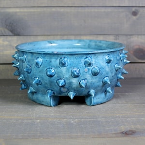 May include: A blue ceramic planter with a spiky, textured exterior and four short legs. The planter has a speckled glaze and a wide, round opening.