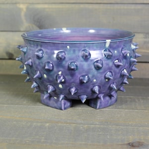 May include: A handcrafted, purple ceramic bowl with a textured surface featuring numerous conical spikes. The bowl has a wide opening and rests on three small feet. The interior has a lighter blue hue.