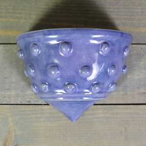 May include: A lavender ceramic wall pocket with a textured surface. The bowl-shaped pocket features a series of raised, rounded bumps around the exterior. The bottom tapers to a point. The wall pocket is designed to be mounted on a wall.
