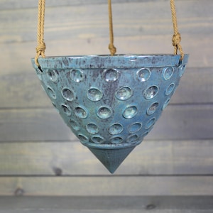 May include: A cone-shaped ceramic hanging planter with a textured blue glaze featuring a repeating pattern of circular indentations. The planter is suspended by three tan-colored twisted rope cords attached to the rim at evenly spaced intervals.