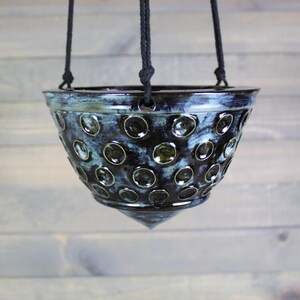May include: A hanging ceramic planter with a dark blue and green glaze. The planter has a series of circular indentations and is suspended by a black rope. The planter has a conical bottom.