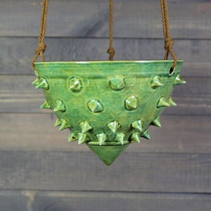 May include: A hanging green ceramic planter with a textured, spiky design. The planter is suspended by brown rope and has a unique, handcrafted appearance. The planter is designed to hold plants and add a decorative touch to any space.