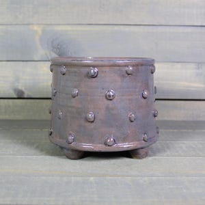 May include: A handcrafted, ceramic planter with a dark, mottled finish. The cylindrical pot features a series of raised, rounded embellishments and rests on three small feet. The planter is approximately 5 inches in diameter and 4 inches tall.