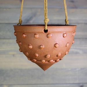 May include: A brown ceramic hanging planter with a textured surface featuring raised, rounded details. The planter has a conical base and is suspended by natural rope. The design is suitable for indoor or outdoor use.