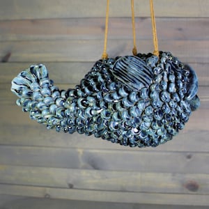 May include: A blue ceramic hanging planter shaped like a fish with a textured, scaly surface. The planter is suspended by a brown twine cord.