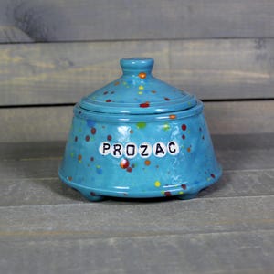 May include: A turquoise ceramic jar with a lid, speckled with colorful dots. The word "PROZAC" is spelled out in white letters on the front of the jar. The jar has a rounded shape and sits on three small feet.