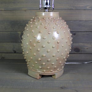 May include: A beige ceramic table lamp with a textured, dotted surface. The lamp has a rounded body and sits on a square base. The lamp has a silver lamp holder and a power cord.