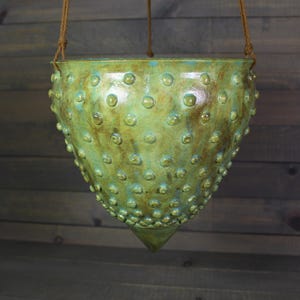 May include: A hanging ceramic planter with a textured surface and a green and brown glaze. The planter has a conical shape with a pointed bottom and is suspended by a brown rope.