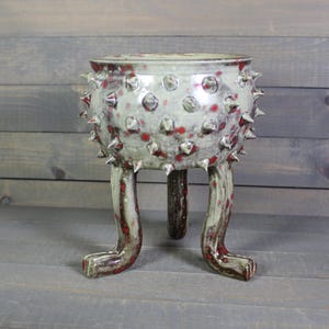 May include: A ceramic planter with a rounded body featuring numerous protruding spikes and a mottled light gray glaze with scattered red spots. The vessel is supported by three distinct, curved legs that extend outward from the base.