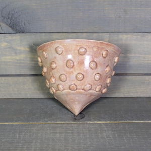 May include: A handmade, light brown ceramic wall pocket with a textured surface of raised, circular bumps. The vessel has a wide, open top and tapers to a pointed bottom. The background is a gray wooden surface.