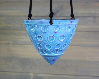 May include: A hanging ceramic planter in a cone shape, glazed in a speckled blue with purple accents. The planter features a pattern of circular indentations and is suspended by three black ropes, ideal for displaying plants.