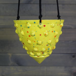May include: A yellow ceramic hanging planter with a speckled design of red, blue, and orange dots. The planter has a textured surface with rounded bumps and is suspended by black rope.
