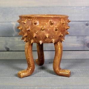 May include: A brown ceramic planter with a textured surface and spiky protrusions. The planter is supported by three curved legs with paw-like feet. The planter has a unique, handcrafted appearance.