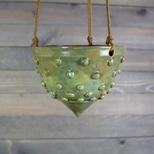 May include: A green ceramic hanging planter with a textured, dotted surface. The planter has a pointed bottom and a hole for drainage.