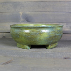 May include: A handcrafted ceramic bowl with a green and blue mottled glaze. The bowl has a rounded shape and sits on three small, square feet. The interior of the bowl is a lighter shade of green.