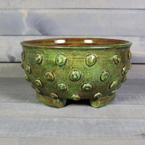 May include: A handcrafted, green ceramic bowl with a textured, dotted surface. The bowl has a rounded shape, sits on three small feet, and features a brown interior. This decorative bowl is suitable for various uses.