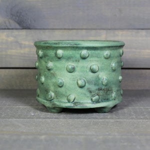 May include: A cylindrical ceramic planter with a sage green glaze and a textured surface featuring raised, evenly spaced circular bumps. The planter has a wide, open top and is supported by three small, integrated feet at the base.