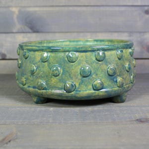May include: A handcrafted ceramic bowl in shades of teal and yellow-green. The bowl features a series of raised, rounded dots around its exterior and rests on three small feet. The interior is a darker teal, and the bowl has a glossy finish.