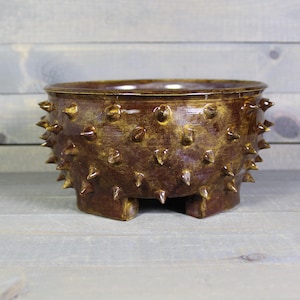 May include: A round ceramic bowl with a glossy brown glaze and a textured surface covered in numerous small, pointed spikes. The bowl features a wide, open top and is supported by short, sturdy feet at the base, creating a distinct, tactile appearance.