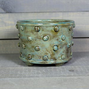 May include: A cylindrical ceramic planter featuring a mottled teal and brown glaze with a raised polka dot texture across the exterior surface. The pot is supported by three small feet at the base and has a smooth, rounded rim at the top.