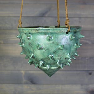 May include: A hanging ceramic planter with a pointed bottom and a textured, spiked surface. The glaze is a mottled seafoam green with dark brown accents. It is suspended by three thick, tan-colored twisted ropes attached to the rim.