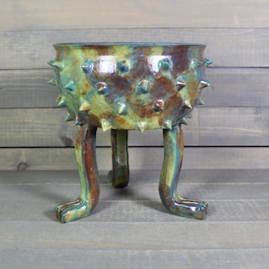May include: A handcrafted ceramic planter with a unique design. The planter features a round bowl with spiky protrusions and is supported by three leg-like structures. The color palette includes shades of green, blue, and brown, creating a textured, artistic appearance.