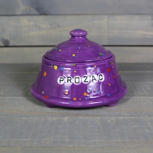 May include: A purple ceramic jar with a lid, featuring the word "PROZAC" in white letters. The jar is decorated with colorful paint splatters. The jar has a rounded shape and sits on three small feet.