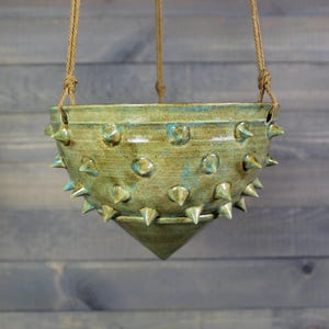 May include: A hanging ceramic planter with a conical base and a glazed green finish. The exterior surface is decorated with numerous protruding spikes arranged in rows. The bowl is suspended by three thick tan ropes attached to the rim.