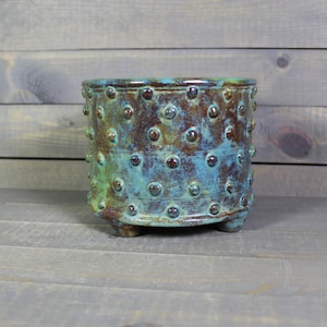 May include: A handcrafted ceramic planter with a textured surface. The planter features a turquoise and brown glaze with raised, rounded accents. It has a cylindrical shape and rests on three small feet, suitable for indoor or outdoor use.