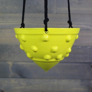 May include: A bright yellow ceramic hanging planter with a textured, dotted surface. The planter has a wide, rounded top and a pointed bottom.