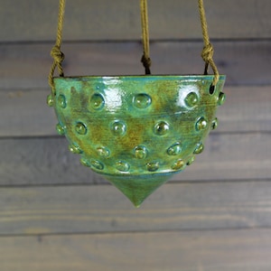 May include: A hanging ceramic planter with a textured, bubble-like design. The planter is glazed in shades of green and turquoise, with a pointed bottom. It is suspended by a natural rope.